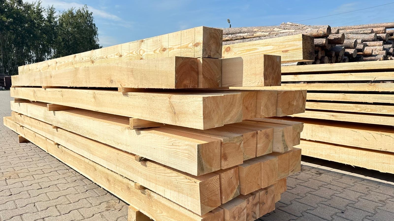 Stos jasnych, heblowanych belek drewnianych ułożonych na palecie, widoczne słoje drewna, w tle składowane pnie drzew