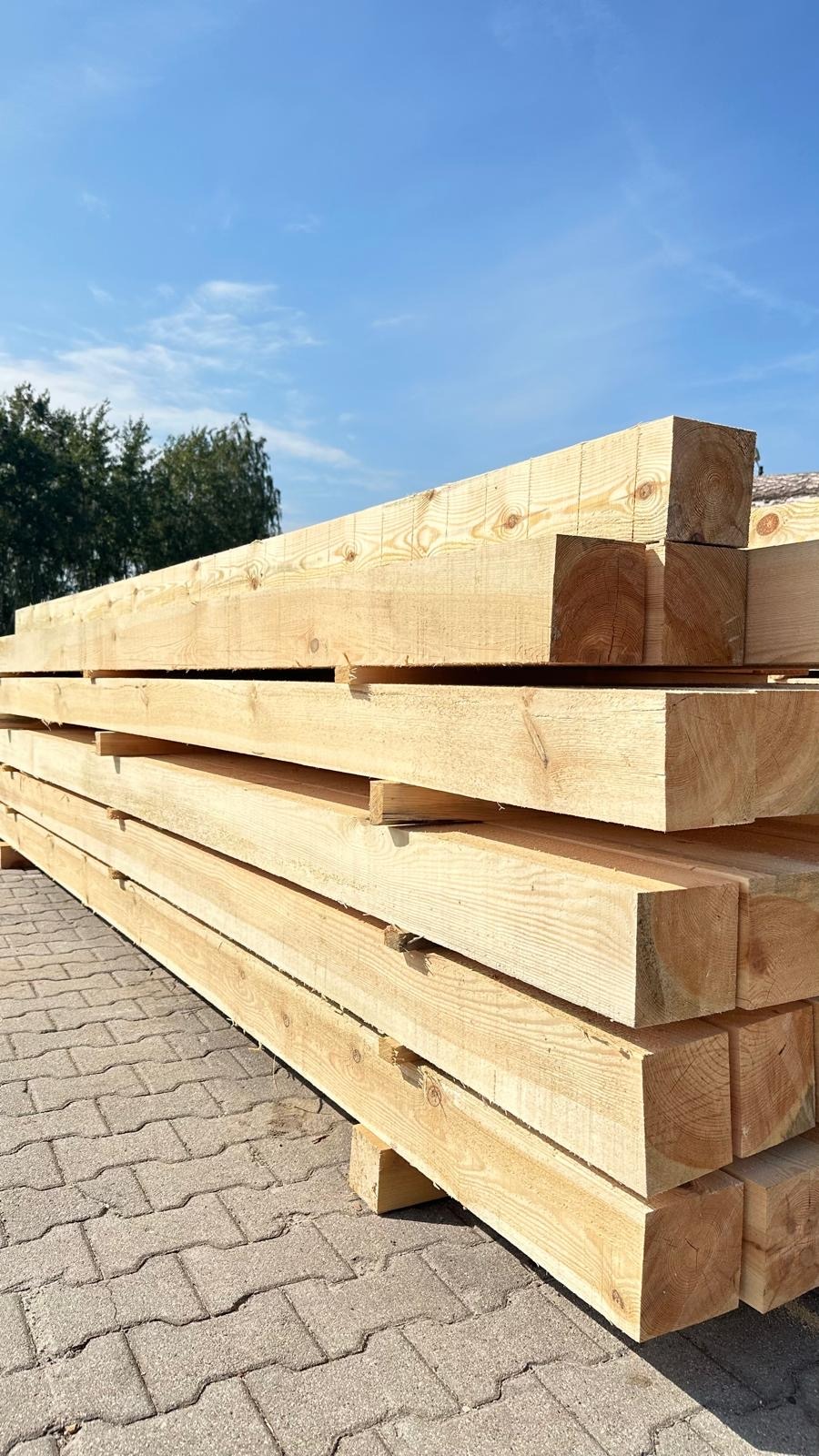 Stos jasnej, świeżo obrobionej tarcicy ułożony na kostce brukowej, widoczne słoje drewna i błękitne niebo w tle.