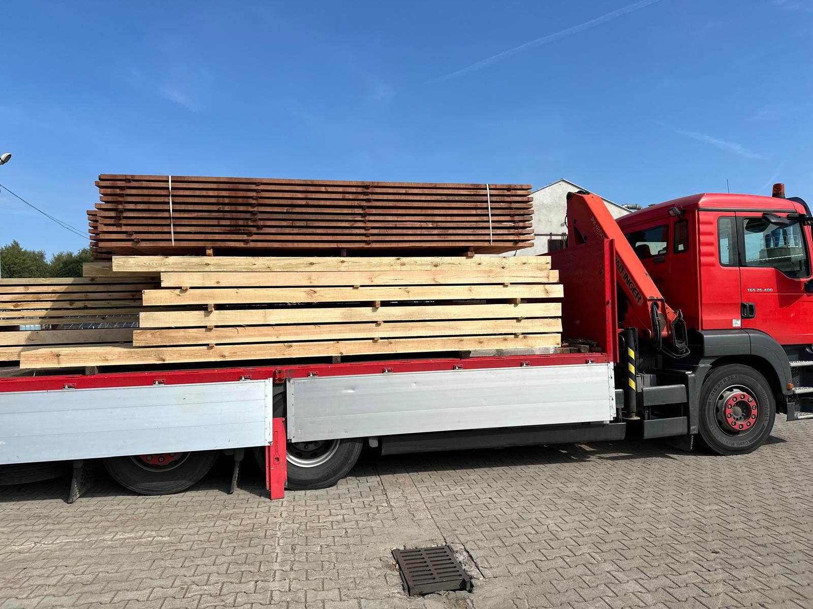 Ciężarówka z dźwigiem Palfinger przewozi ładunek tarcicy w Szczytkowicach. Na pace ułożone są dwie warstwy drewna: jasne, surowe deski oraz ciemniejsze, prawdopodobnie impregnowane. Błękitne niebo...