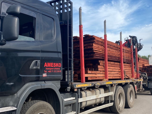 Ciężarówka z czarną kabiną marki Scania, załadowana równo ułożonymi deskami z drewna, zabezpieczonymi czerwonymi słupkami, widoczne logo firmy transportowej AMESKO