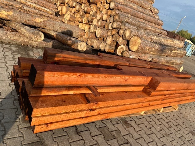 Stos belek z obrobionego drewna na palecie, w tle pryzma okrąglaków i kostka brukowa.