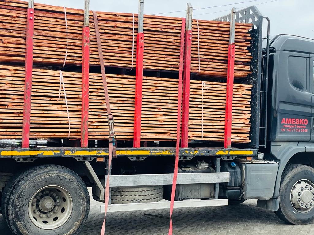 Ciężarówka z załadowanym stosem tarcicy budowlanej zabezpieczonej czerwonymi pasami transportowymi, widoczne opony i błotniki pojazdu.