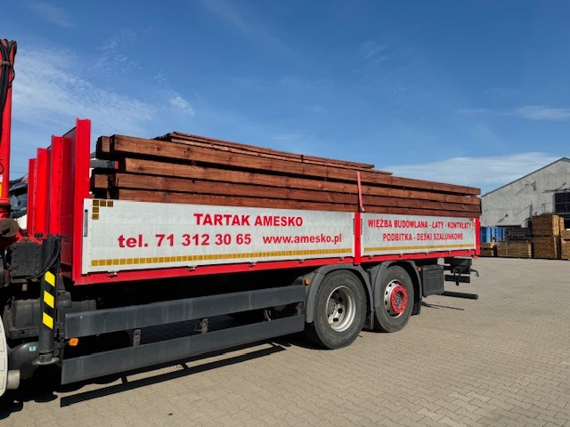 Ciężarówka z czerwonym nadwoziem, załadowana brązową tarcicą budowlaną, z widocznym logo tartaku Amesko i numerem telefonu na boku, na tle błękitnego nieba i hali magazynowej.