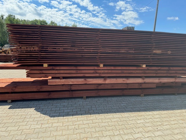 Stosy impregnowanej tarcicy w kolorze ciemnego brązu, ułożone na placu z kostki brukowej, widoczne niebo i drzewa w tle.