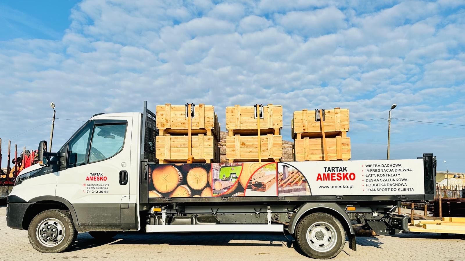 Ciężarówka z logo Tartak Amesko przewozi skrzynie z drewnem, widoczne także grafiki z przekrojami drewna i listą usług: więźba dachowa, impregnacja drewna, łaty, deska szalunkowa, podbitka dachowa,...