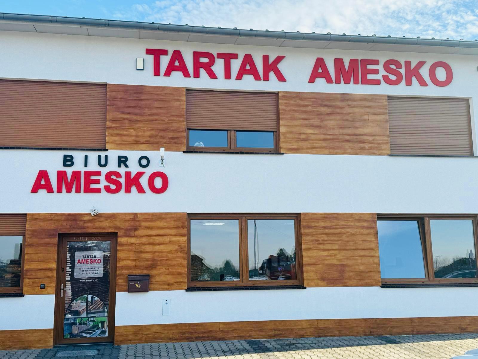Fasada budynku z napisem 'TARTAK AMESKO' i 'BIURO AMESKO' w czerwonych literach, obłożona panelami imitującymi drewno, z brązowymi oknami i drzwiami wejściowymi z naklejką informacyjną.