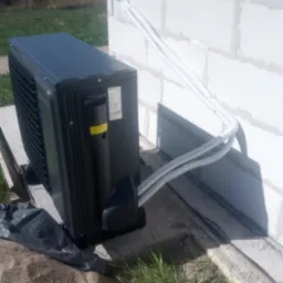 Czarna jednostka zewnętrzna pompy ciepła Seyva zamontowana przy ścianie budynku z betonu komórkowego, z widocznymi przewodami w peszlach.