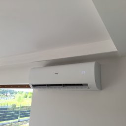 KlimaWentKomfort Mirosław Gawin - Biały klimatyzator Haier zamontowany na jasnej ścianie w pokoju z widokiem na zieleń przez okno. Na wyświetlaczu widoczna temperatura 23 stopnie.