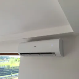 Biały klimatyzator Haier zamontowany na jasnej ścianie w pokoju z widokiem na zieleń przez okno. Na wyświetlaczu widoczna temperatura 23 stopnie.