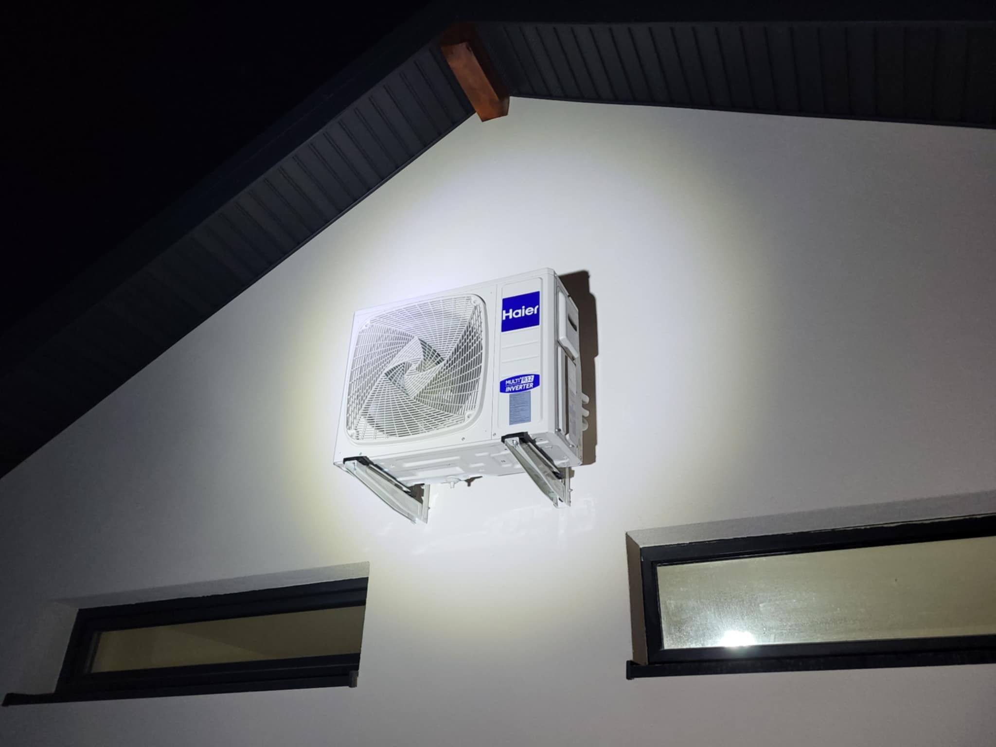 Biała jednostka zewnętrzna klimatyzacji Haier zamontowana na białej ścianie budynku z dwoma oknami, widoczna pod kątem od dołu, oświetlona punktowo.