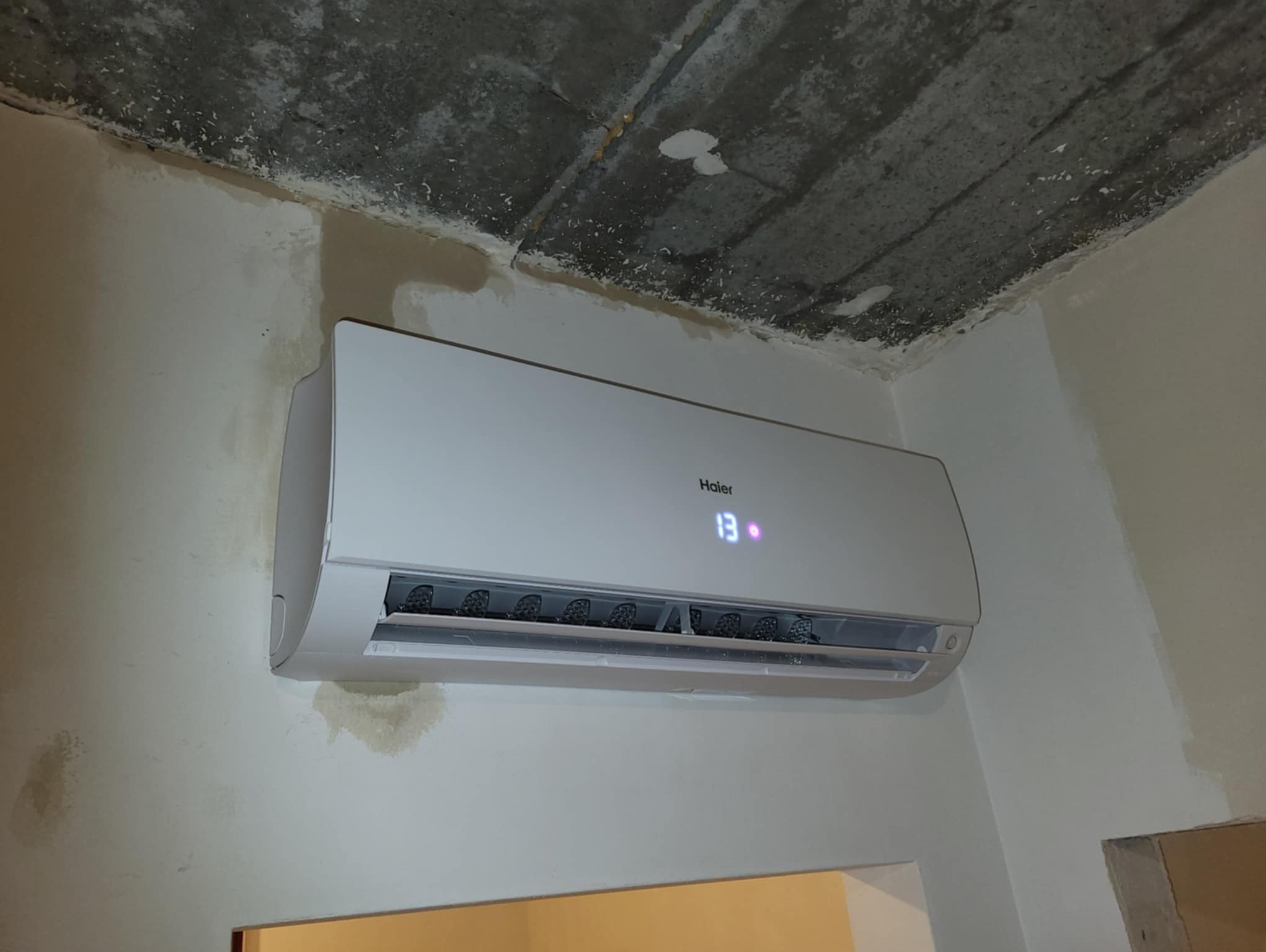 Zamontowany klimatyzator marki Haier z wyświetlaną temperaturą 13 stopni Celsjusza na ścianie w trakcie wykańczania, widoczny surowy sufit.