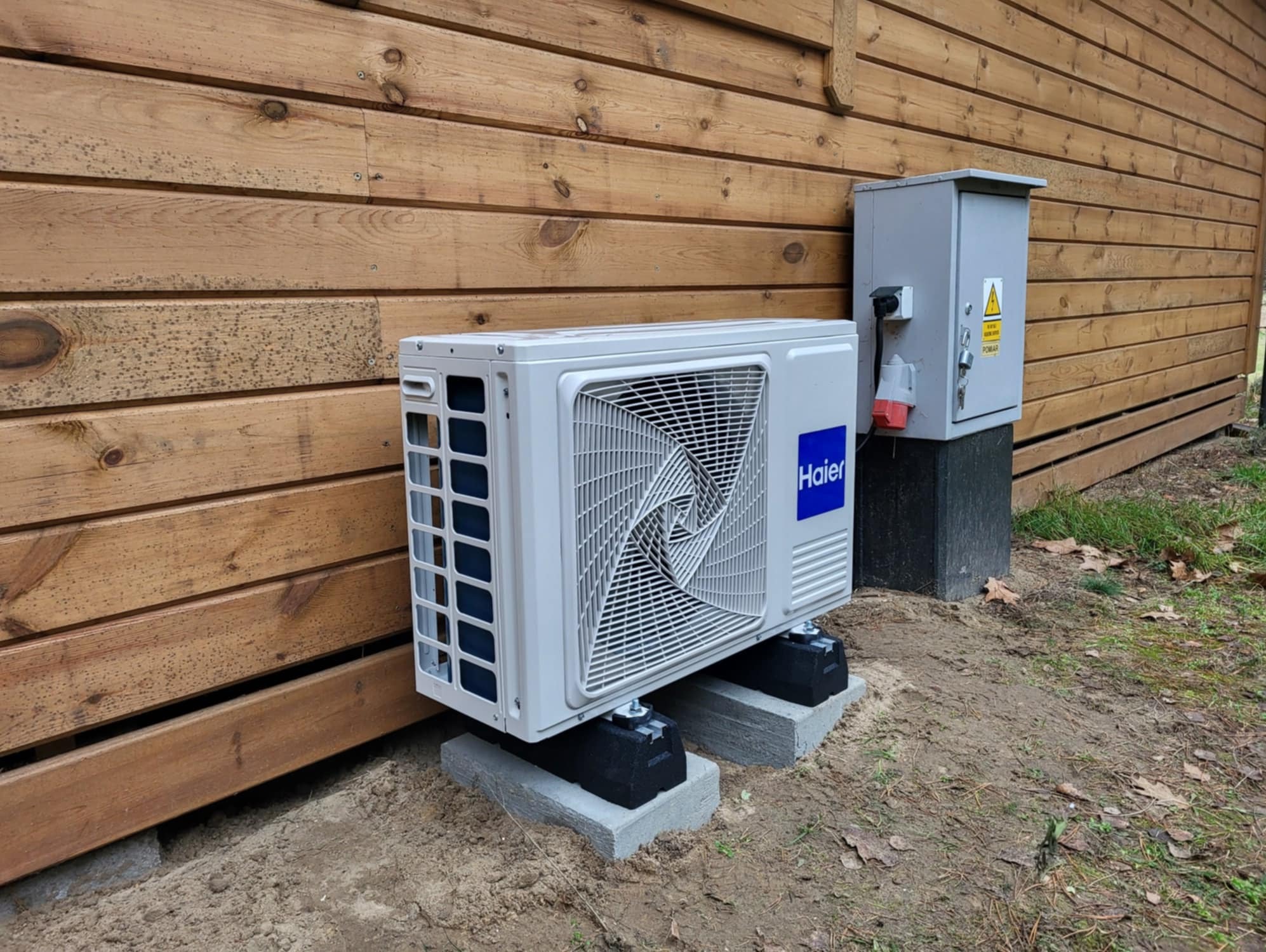 Biała jednostka zewnętrzna klimatyzacji Haier zamontowana na betonowych i gumowych podstawach przy drewnianej ścianie budynku, obok szara skrzynka elektryczna z gniazdem i wtyczką.