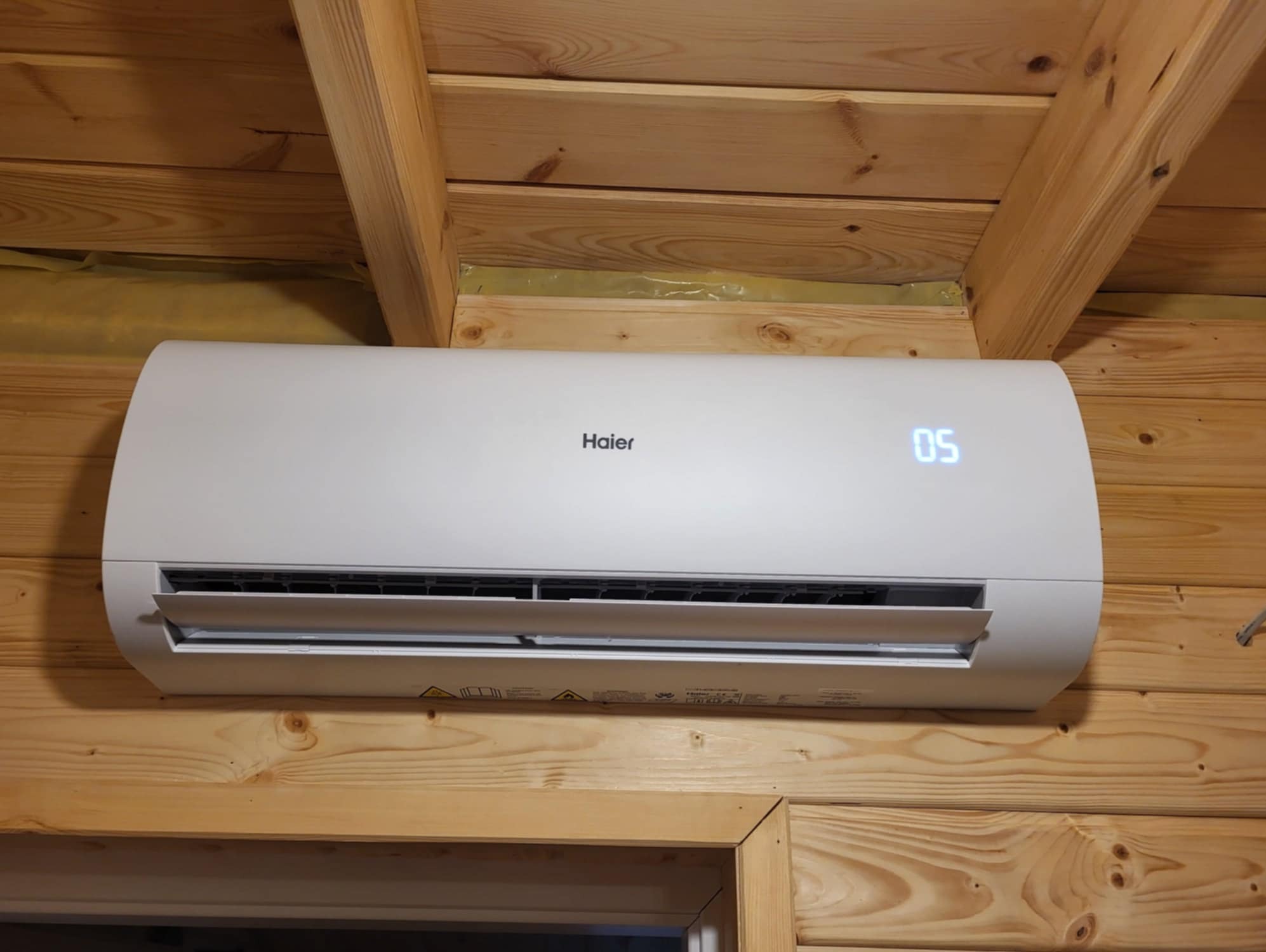 Biały klimatyzator Haier zamontowany na drewnianej ścianie z bali, wyświetlający temperaturę 05.
