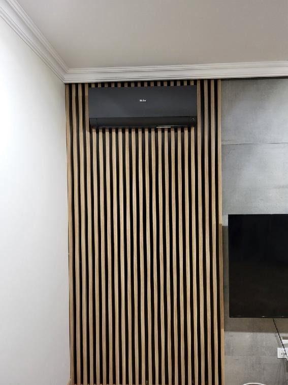 Czarny klimatyzator na ścianie z drewnianymi listwami, obok betonowa ściana z telewizorem, biały sufit z dekoracyjną listwą.