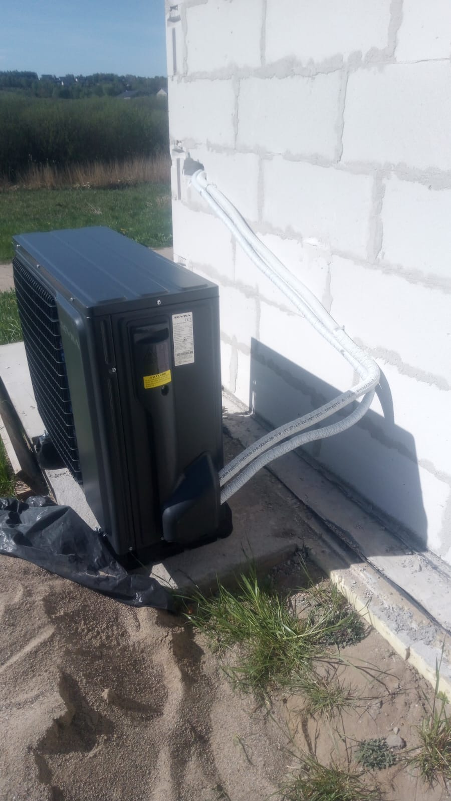 Czarna jednostka zewnętrzna pompy ciepła Seyva zamontowana przy ścianie budynku z betonu komórkowego, z widocznymi przewodami w peszlach.
