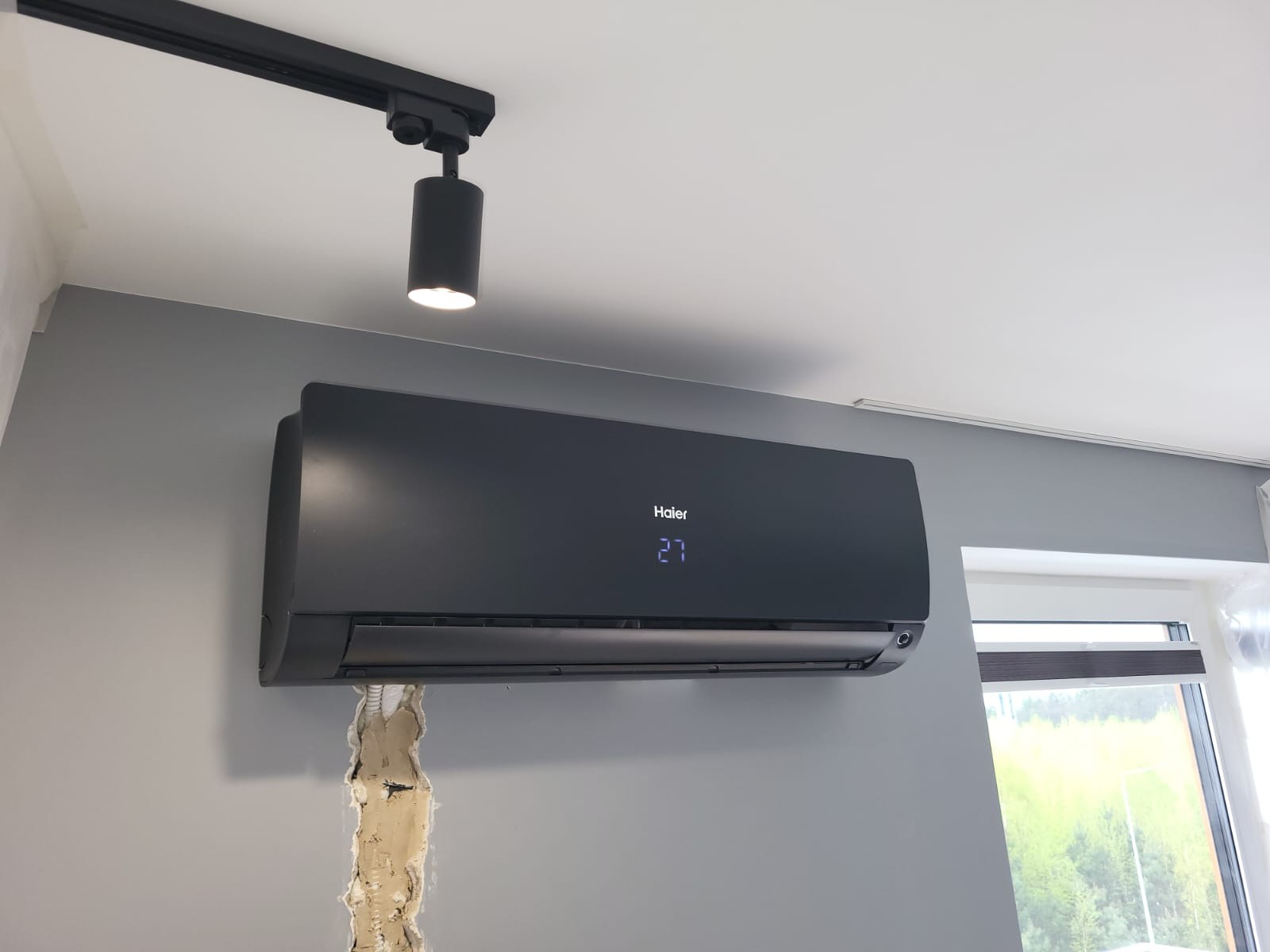 Czarny klimatyzator Haier na szarej ścianie, pod czarnym reflektorem szynowym. Widoczna instalacja elektryczna w ścianie. Okno z roletą w tle.