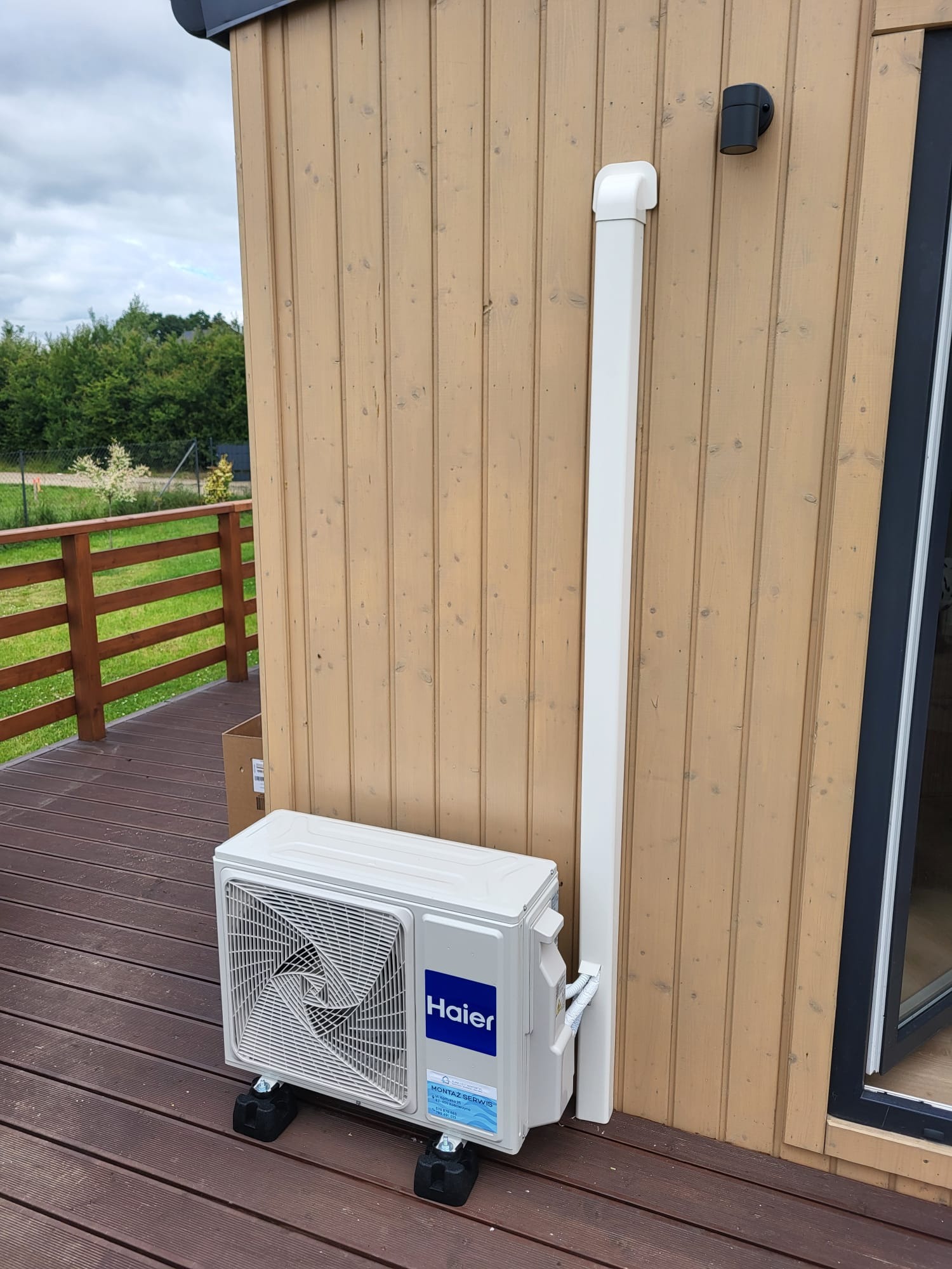 Biała jednostka zewnętrzna klimatyzacji Haier na drewnianym tarasie przy ścianie domu z drewnianą elewacją i białą rurą odprowadzającą skropliny.