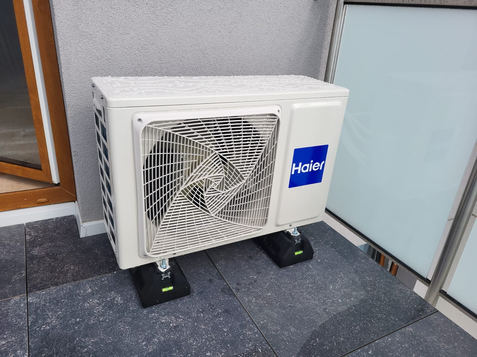 Biała jednostka zewnętrzna klimatyzacji Haier na czarnych podkładkach z poziomicą, ustawiona na szarym tarasie przy ścianie budynku. Widoczne krople wody.
