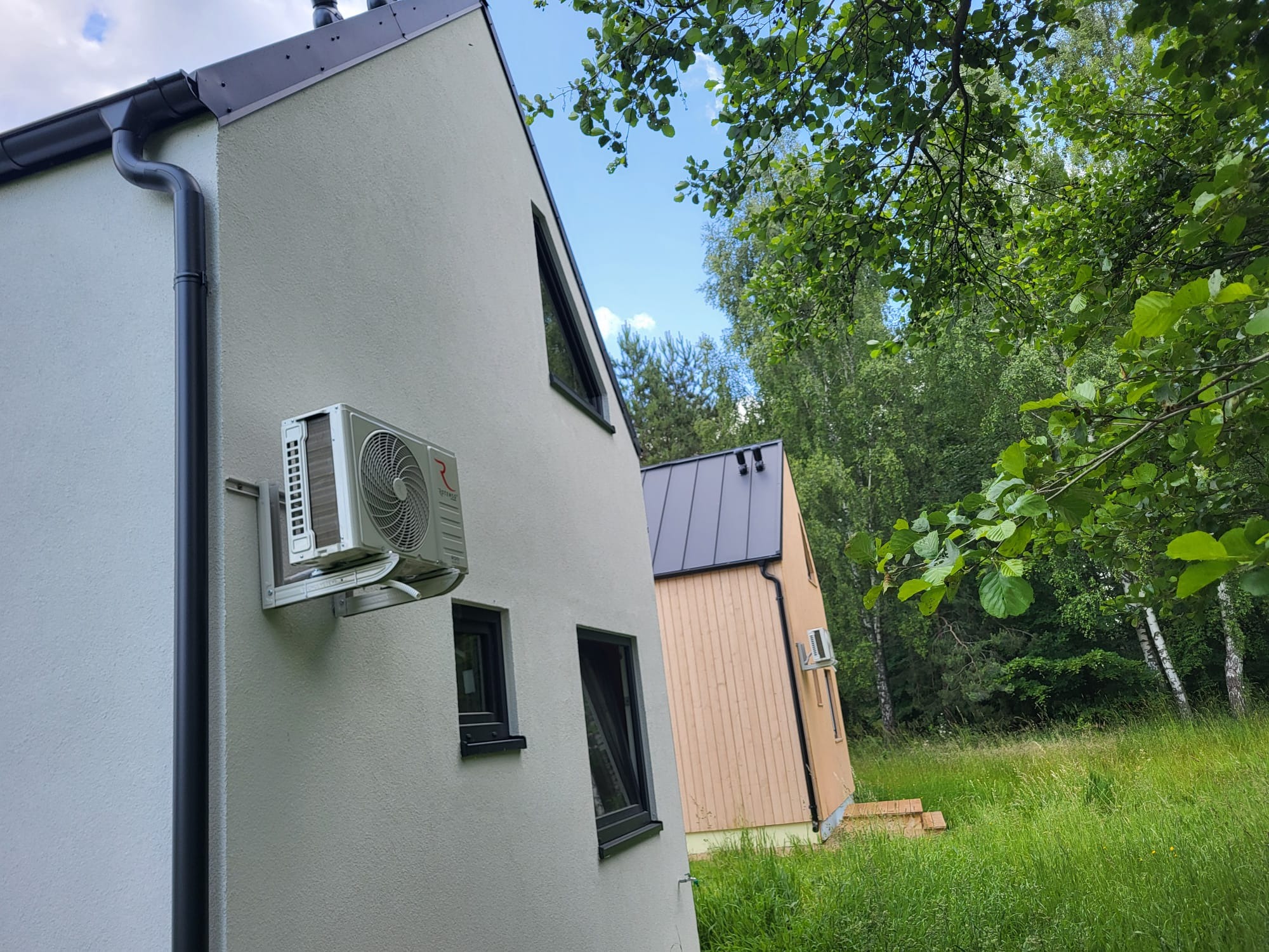 Klimatyzator na elewacji jasnego domu z ciemnymi oknami, w tle drugi dom i zieleń drzew. Widok z dołu, błękitne niebo i gałęzie drzew w górnym rogu.