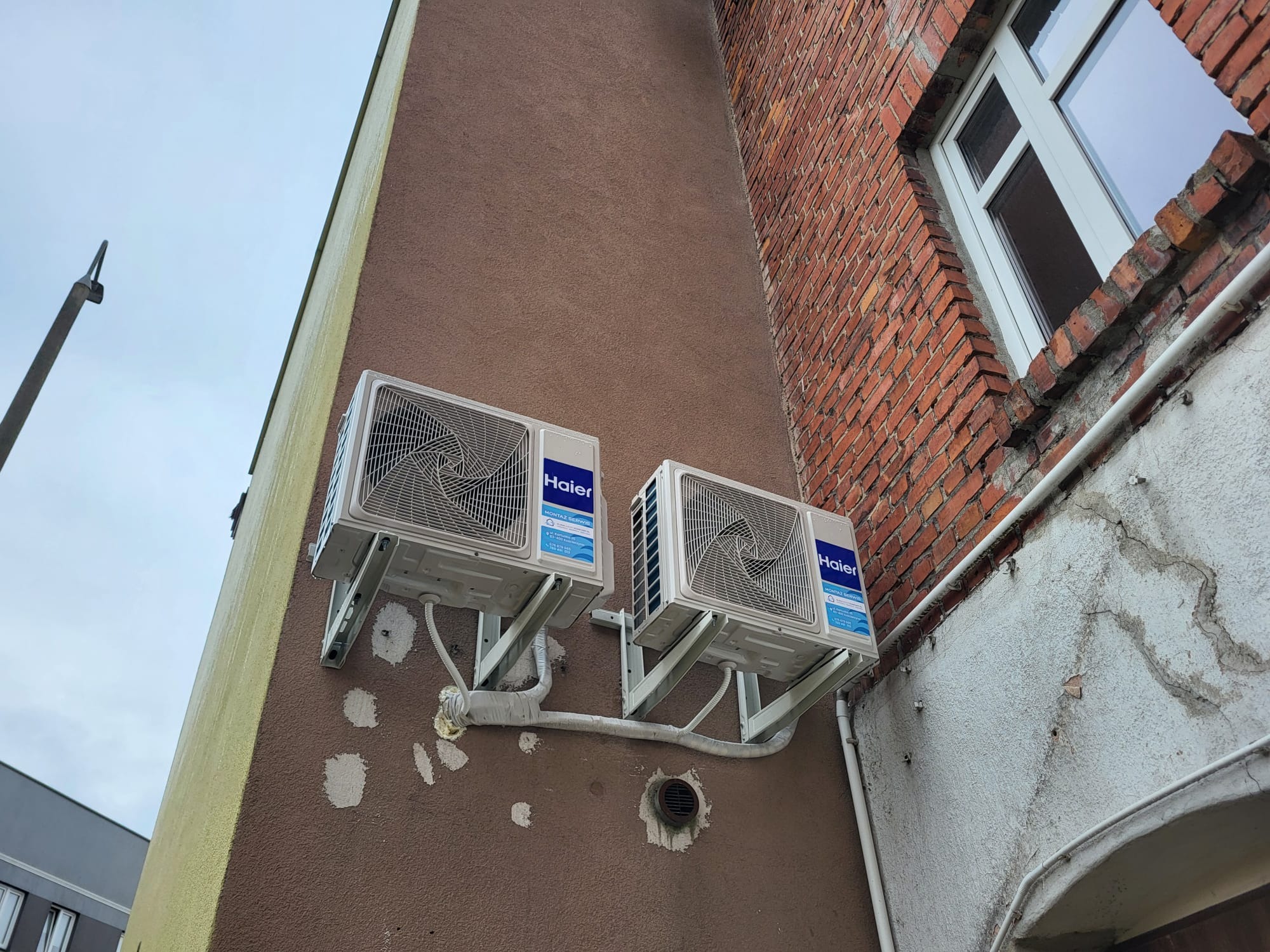 Dwa klimatyzatory Haier zamontowane na ścianie budynku z brązowym tynkiem i ceglaną ścianą. Widoczne białe rury i wsporniki montażowe.