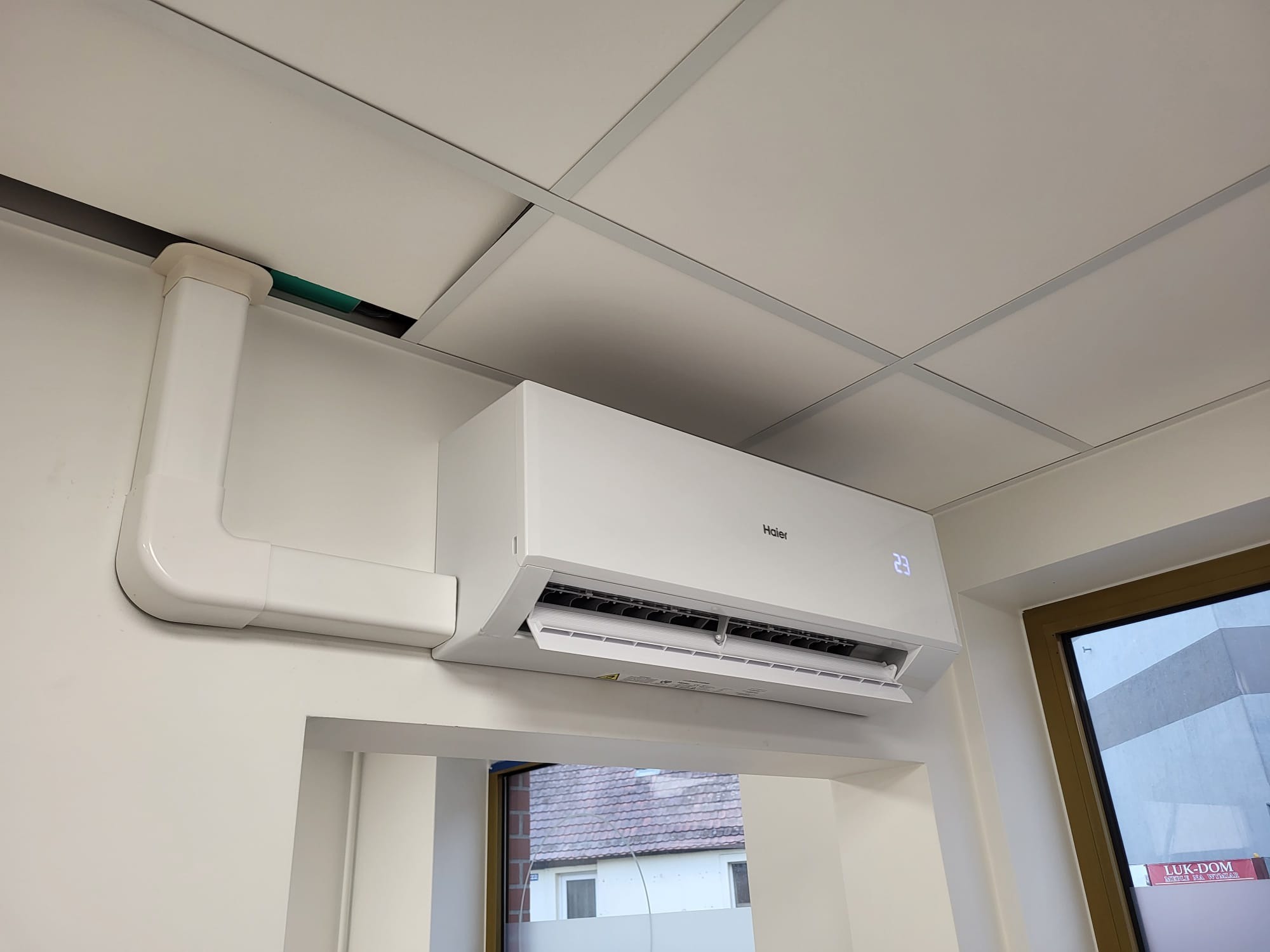 Biały klimatyzator Haier zamontowany na ścianie, z widoczną instalacją prowadzącą do sufitu kasetonowego. Widok z dołu, z oknem w tle.