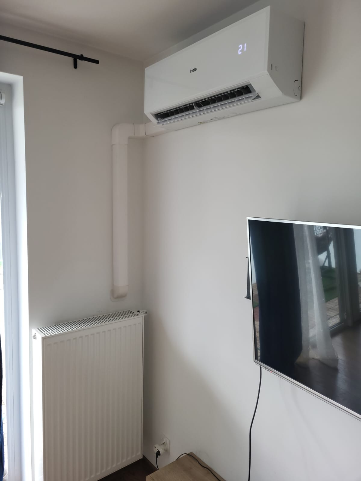Biały klimatyzator Haier zamontowany na ścianie w pokoju, z widoczną instalacją odprowadzającą skropliny. Obok grzejnik i telewizor.