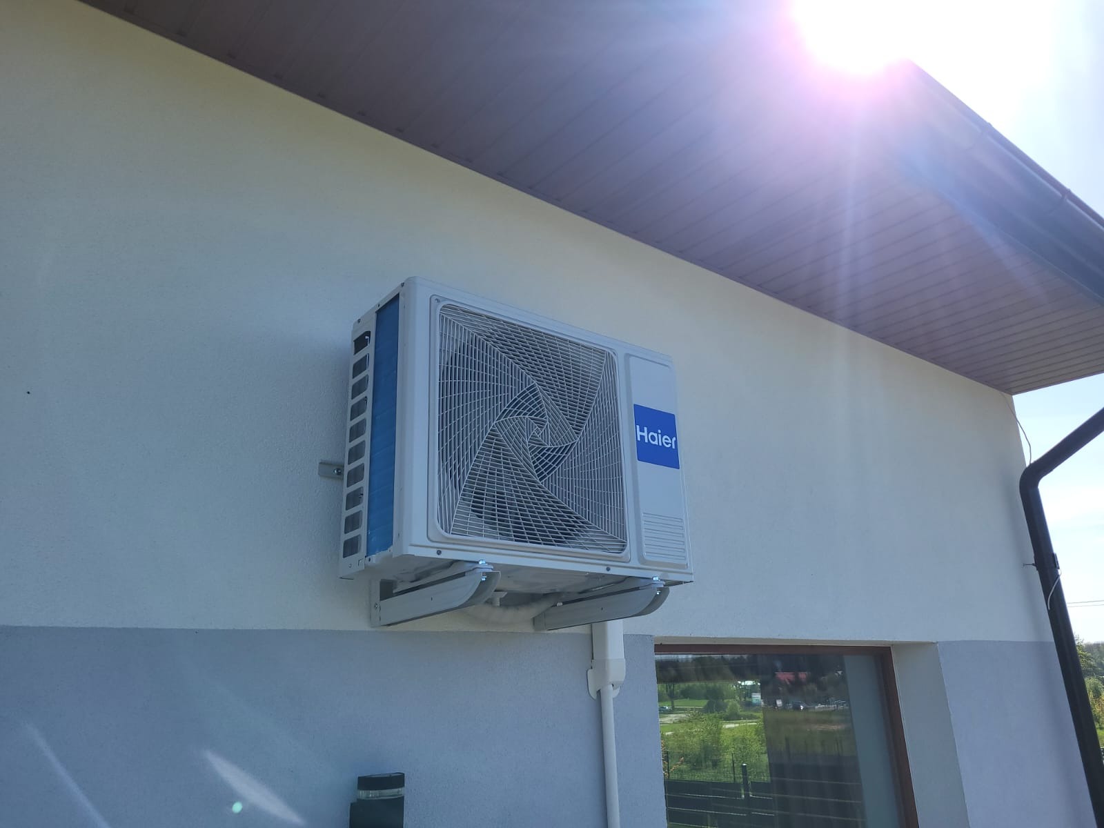 Biała jednostka zewnętrzna klimatyzacji Haier zamontowana na elewacji domu, widok z dołu, jasne światło słoneczne.