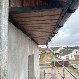 Usługi dekarskie - Montaż okapu dachowego z blachy trapezowej imitującej drewno, widok z rusztowania na surową elewację budynku i zabudowania gospodarcze w tle.