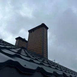 Nowy, czarny dach z blachy trapezowej z dwoma kominami obłożonymi brązową boazerią na tle pochmurnego nieba.