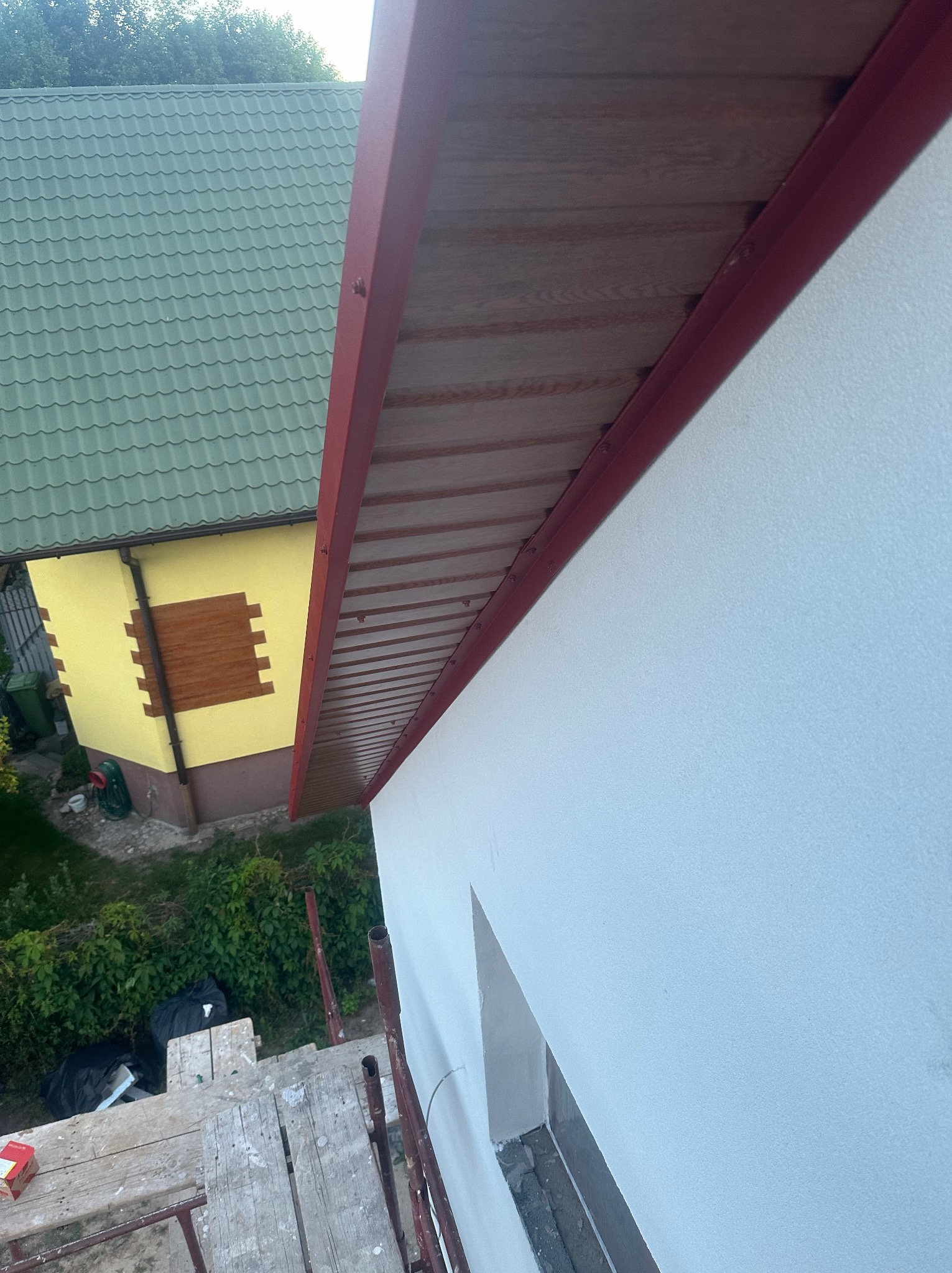 Widok z rusztowania na fragment ściany budynku z widocznym oknem w trakcie prac wykończeniowych, z brązową podbitką i czerwoną listwą okapową, w tle zielony dach sąsiedniego budynku.