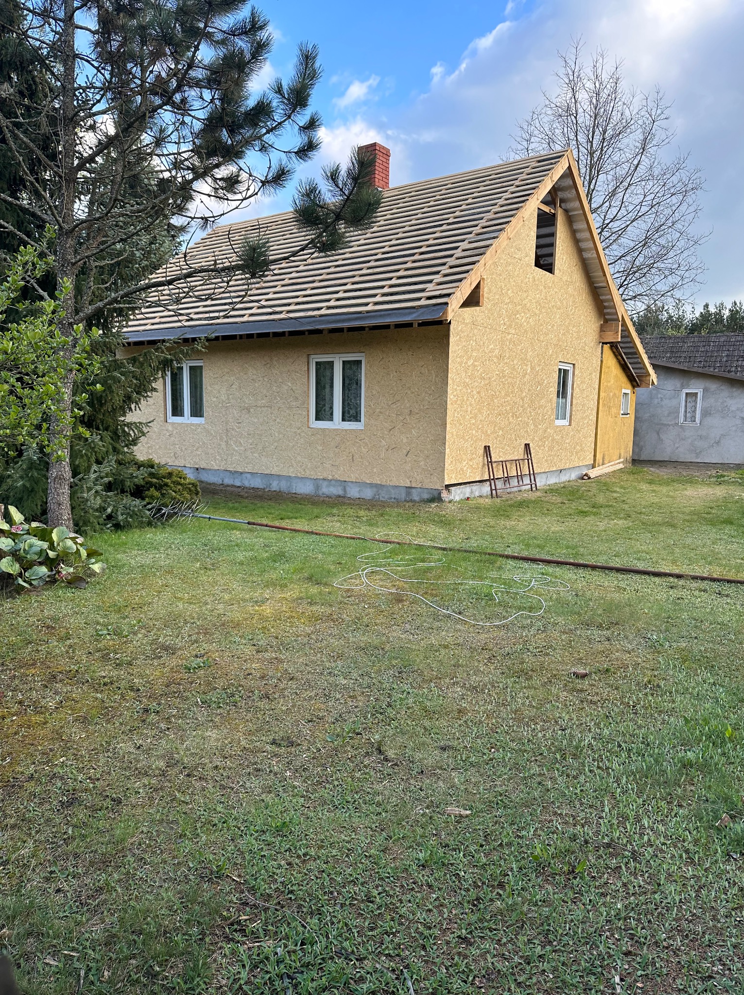 Dom w trakcie budowy z widoczną konstrukcją dachu i płytami OSB na ścianach zewnętrznych, na trawniku leżą węże i rura, a obok domu stoi mała drabinka.