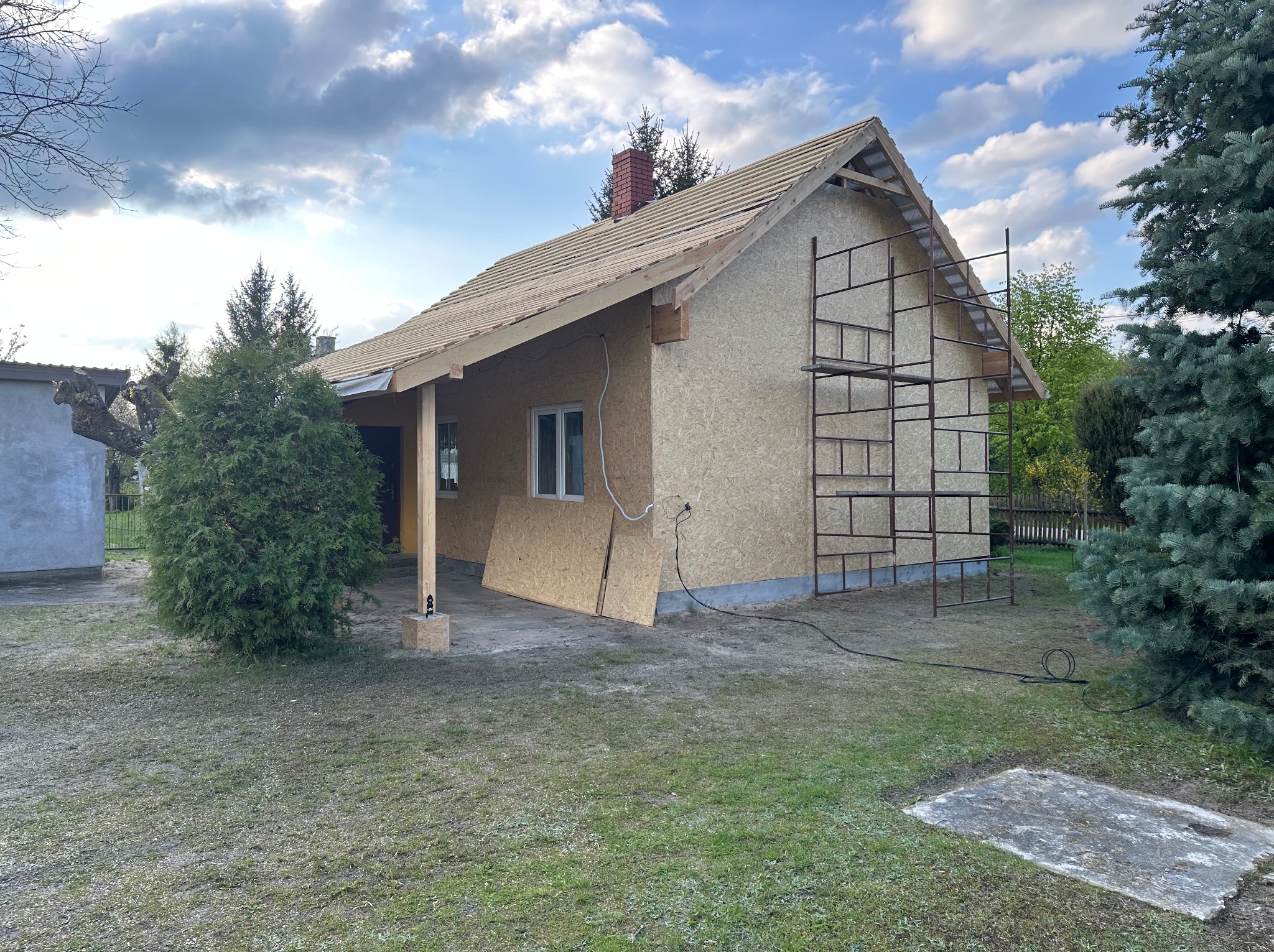 Dom jednorodzinny w trakcie prac dekarskich: przygotowanie podłoża pod nowe pokrycie, widoczne płyty OSB na dachu i ścianach, rusztowanie przy elewacji, kabel zasilający na trawniku.