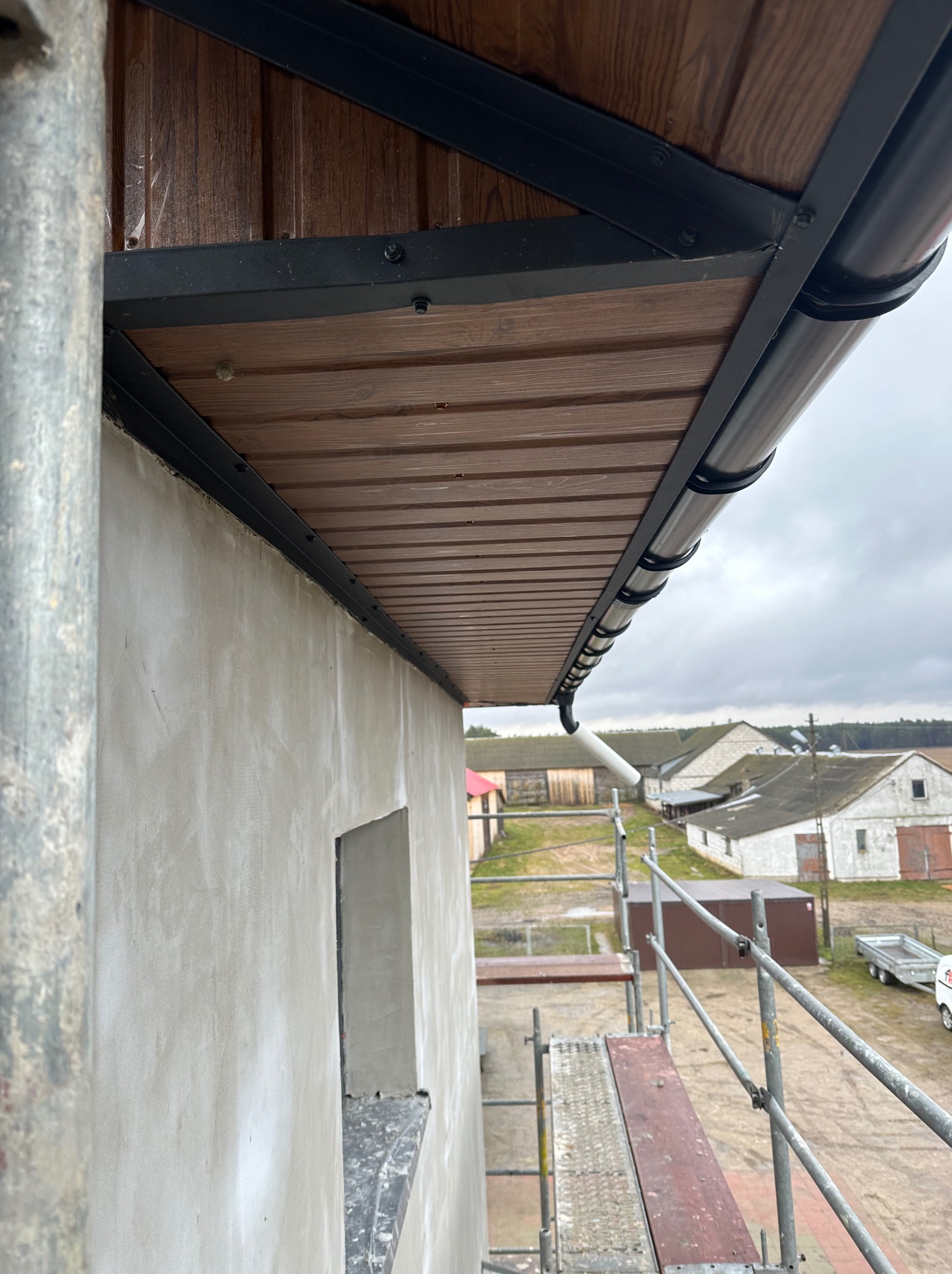 Montaż okapu dachowego z blachy trapezowej imitującej drewno, widok z rusztowania na surową elewację budynku i zabudowania gospodarcze w tle.