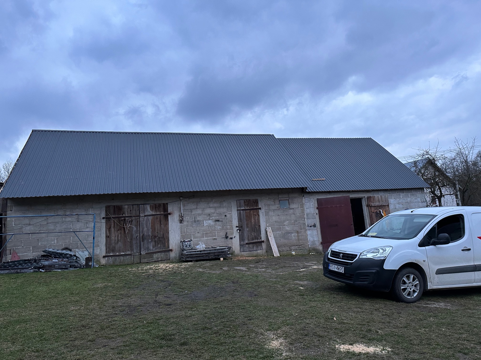 Budynek gospodarczy z nowym, szarym dachem z blachy trapezowej, zaparkowany biały bus dostawczy, pochmurne niebo.