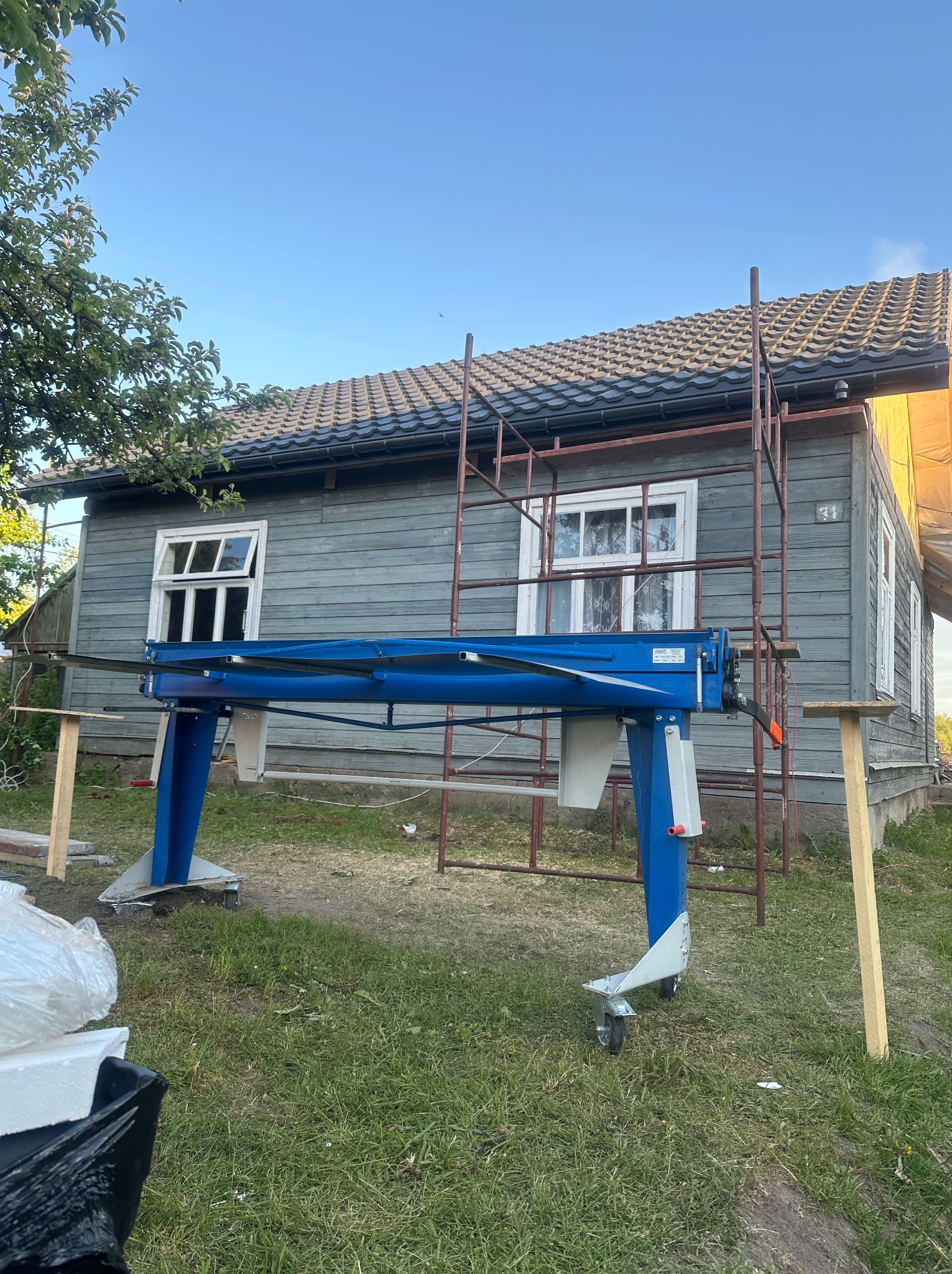Niebieska maszyna do obróbki blachy na tle domu z szarą elewacją i częściowo zdjętym pokryciem, widoczne rusztowanie.