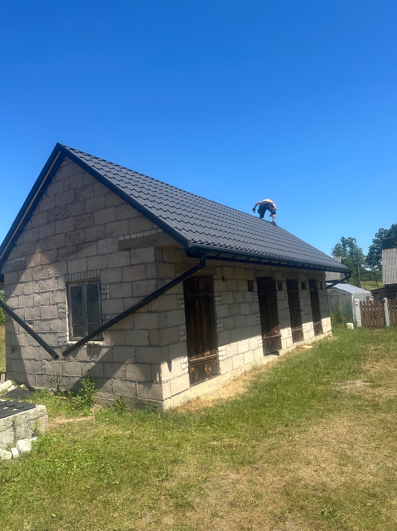 Praca na dachu jednopiętrowego budynku z bloków. Widoczny pracownik na dachu z ciemną, nową dachówką. Budynek ma drewniane okiennice i rynny. W tle błękitne niebo i zieleń.