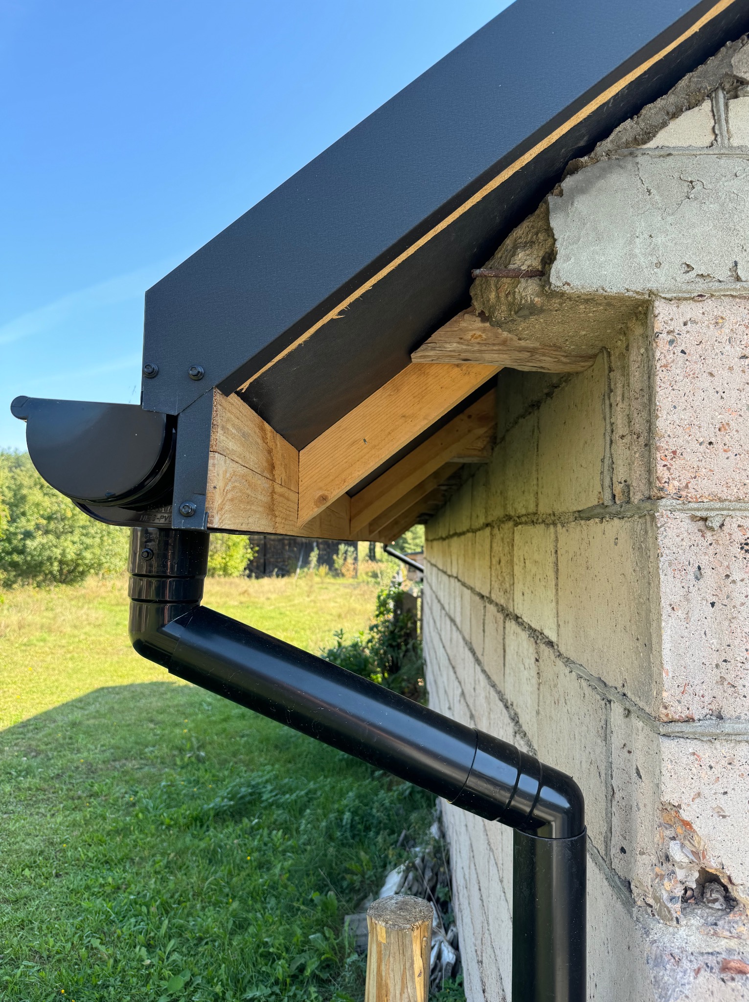 Czarny system rynnowy zamontowany przy ścianie budynku z bloczków betonowych, widoczna drewniana konstrukcja dachu i fragment zielonego trawnika.