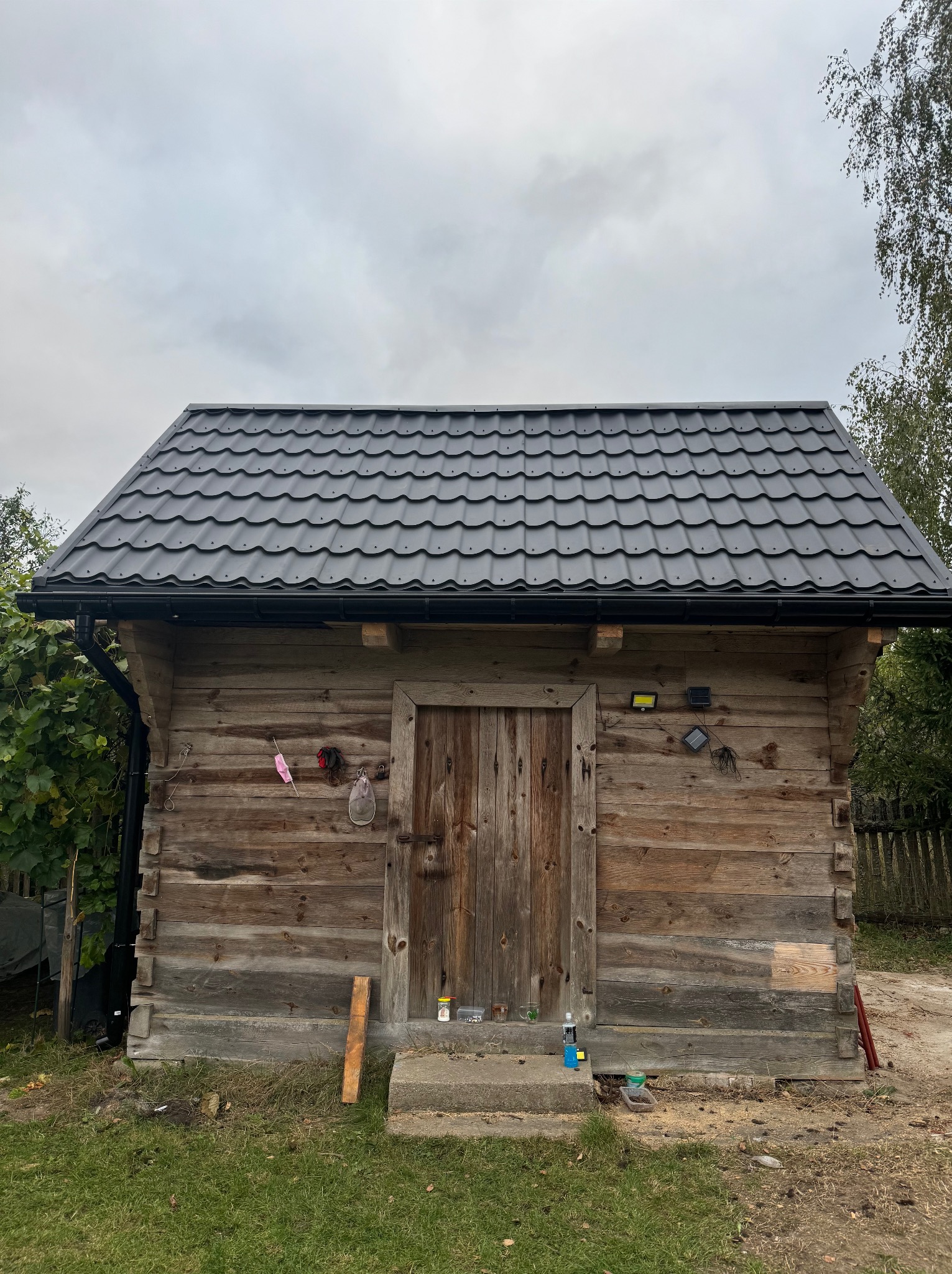 Mała drewniana szopa z nowym, ciemnoszarym dachem z blachy dachówkowej, widoczne obróbki blacharskie, rynny i oświetlenie solarne na ścianie, narzędzia i akcesoria wiszące na elewacji.