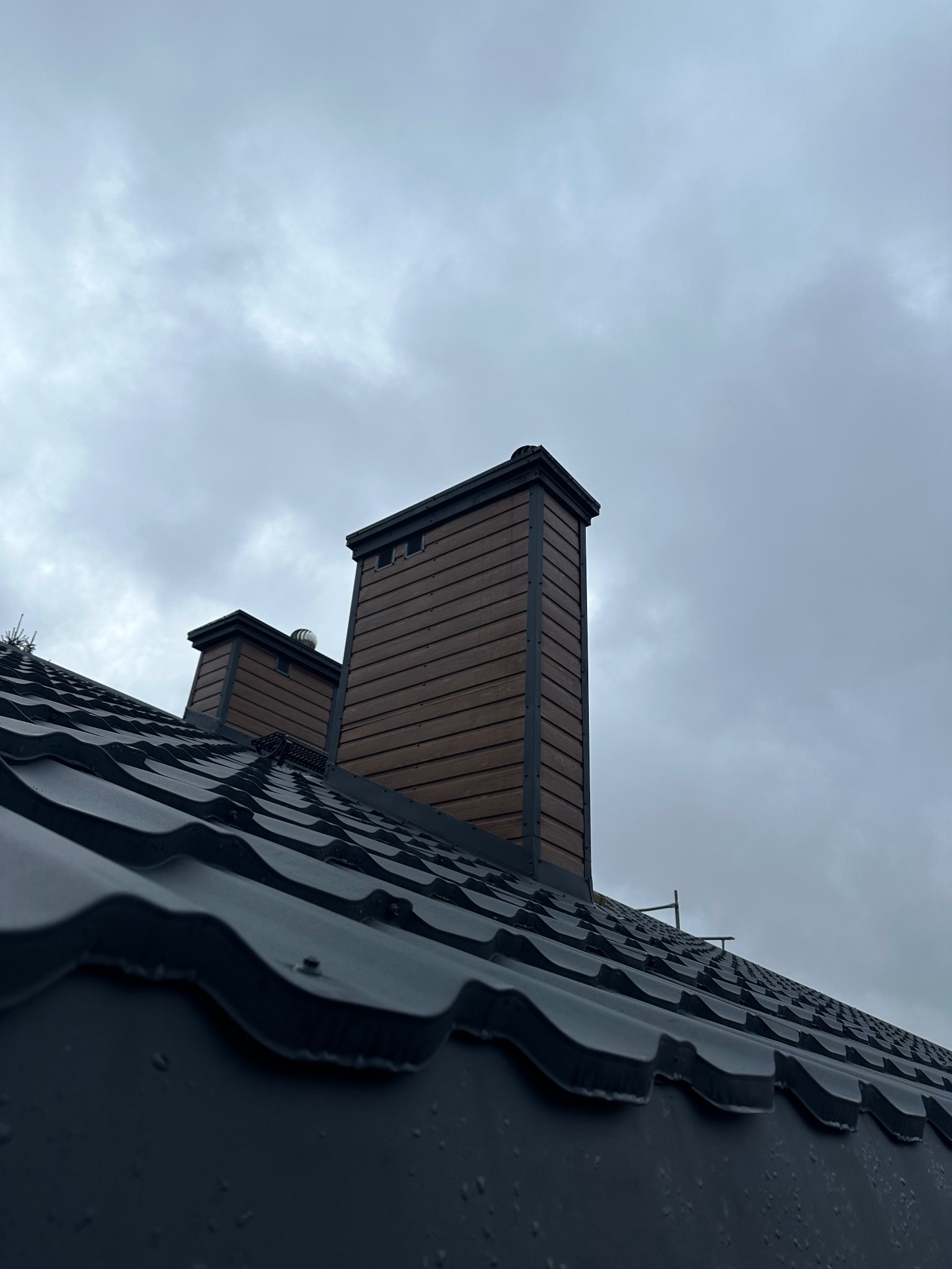 Nowy, czarny dach z blachy trapezowej z dwoma kominami obłożonymi brązową boazerią na tle pochmurnego nieba.