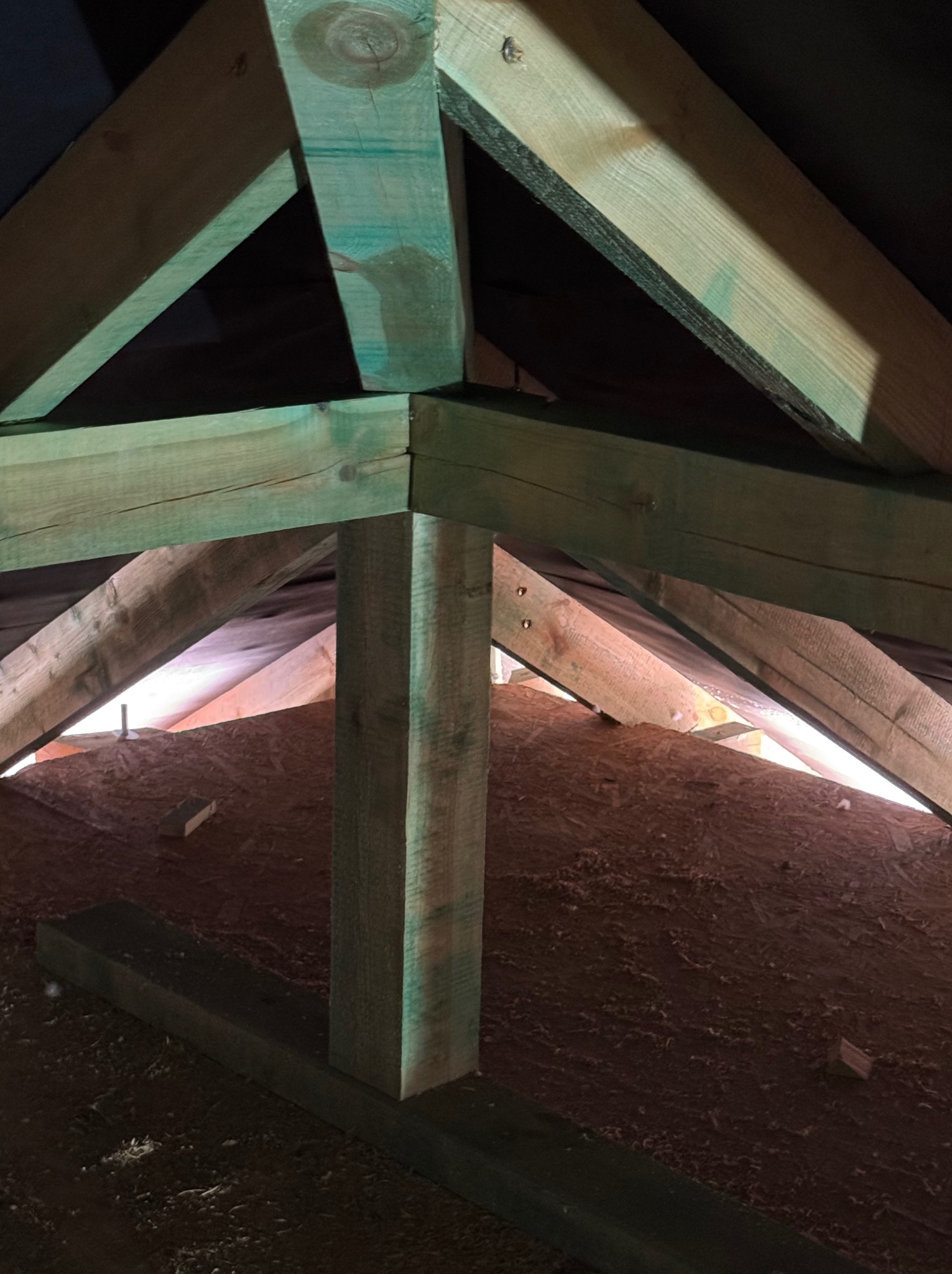 Fragment drewnianej konstrukcji dachu z zielonymi impregnatami, widok z dołu, ukazujący połączenia belek i słupów w surowym stanie.