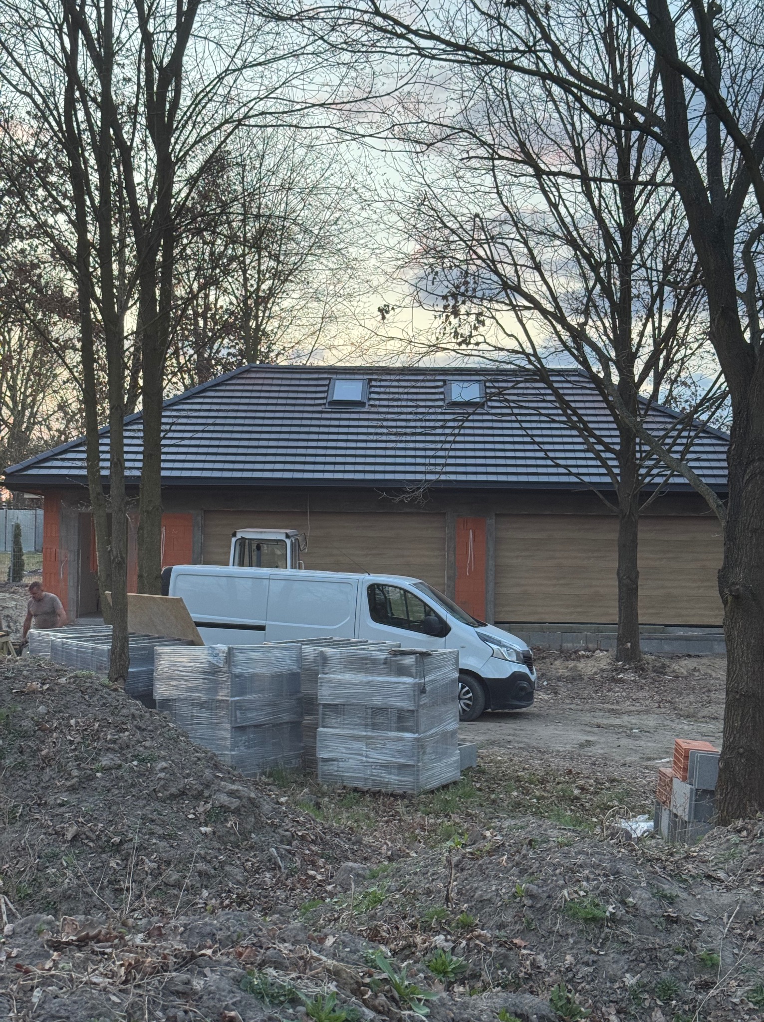Budynek w trakcie budowy z ciemną dachówką, widoczne okna dachowe, zaparkowany biały bus i materiały budowlane. Prace w toku.
