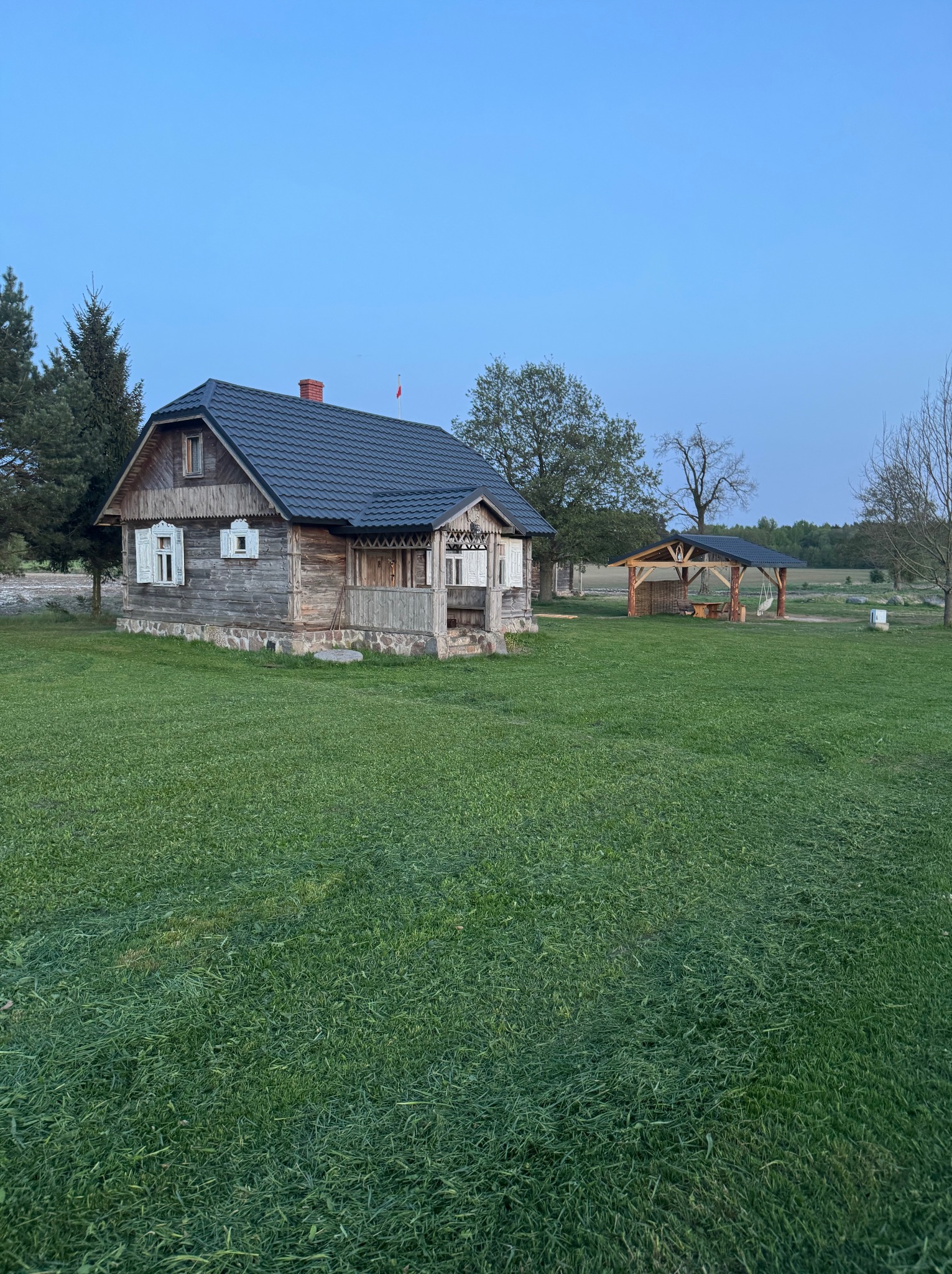 Drewniany dom z ciemnym dachem na zielonej trawie, w tle altana. Ujęcie wczesnym rankiem, miękkie światło podkreśla fakturę drewna i detale architektoniczne.