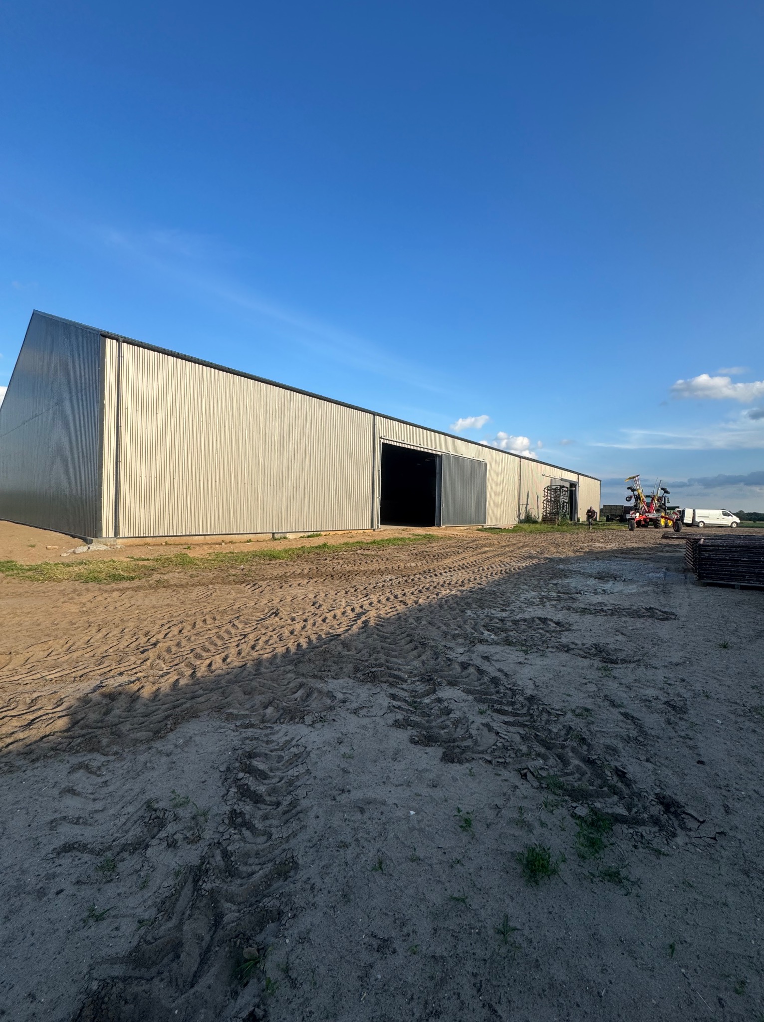Nowoczesna hala stalowa z blachy trapezowej w neutralnych barwach, widoczna z zewnątrz na tle błękitnego nieba. Na pierwszym planie ziemia z wyraźnymi śladami opon.