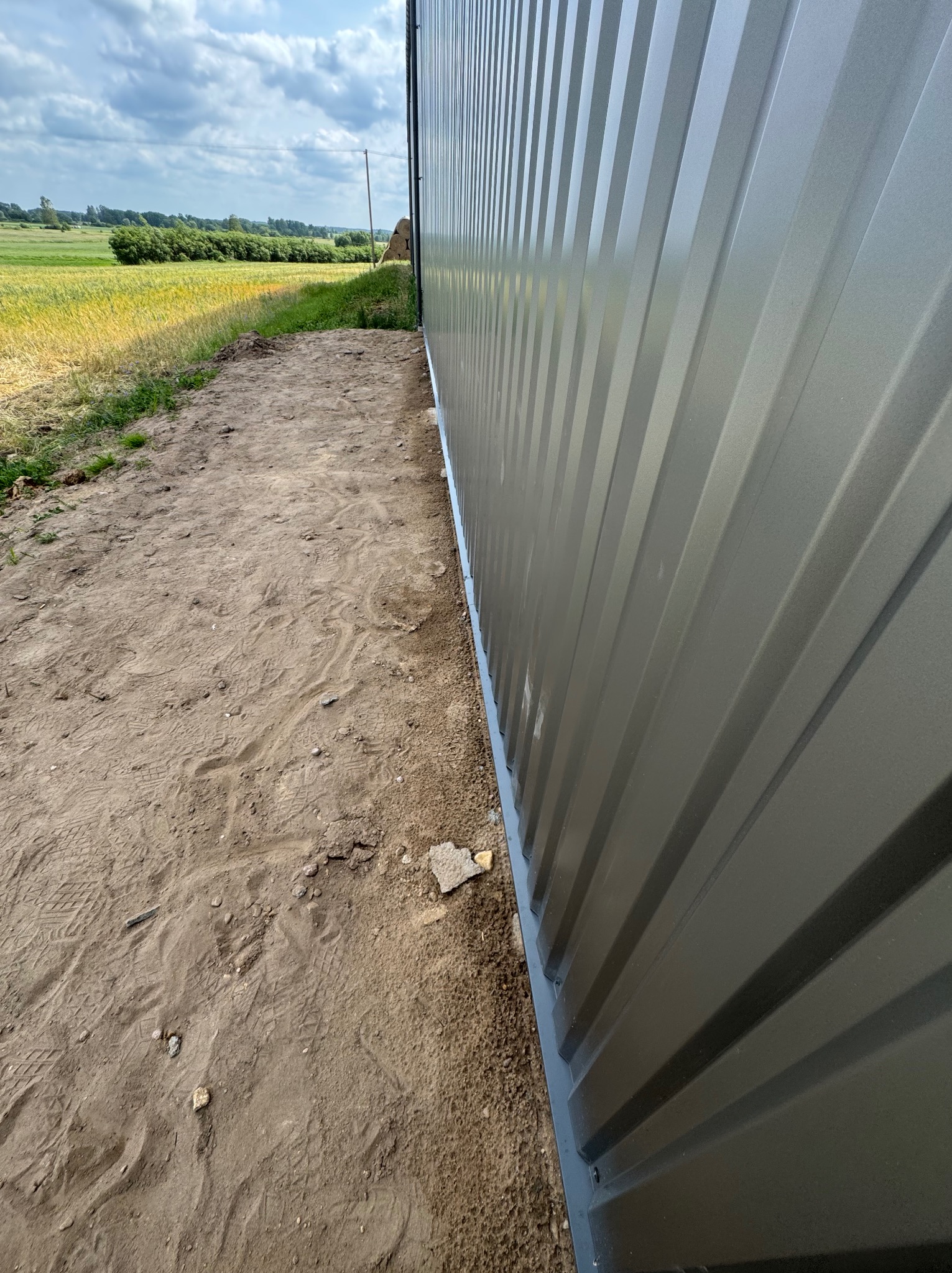 Szara blacha trapezowa tworząca ścianę hali, widoczna z bliska, z polami w tle. Detal konstrukcji hali z blachy trapezowej.