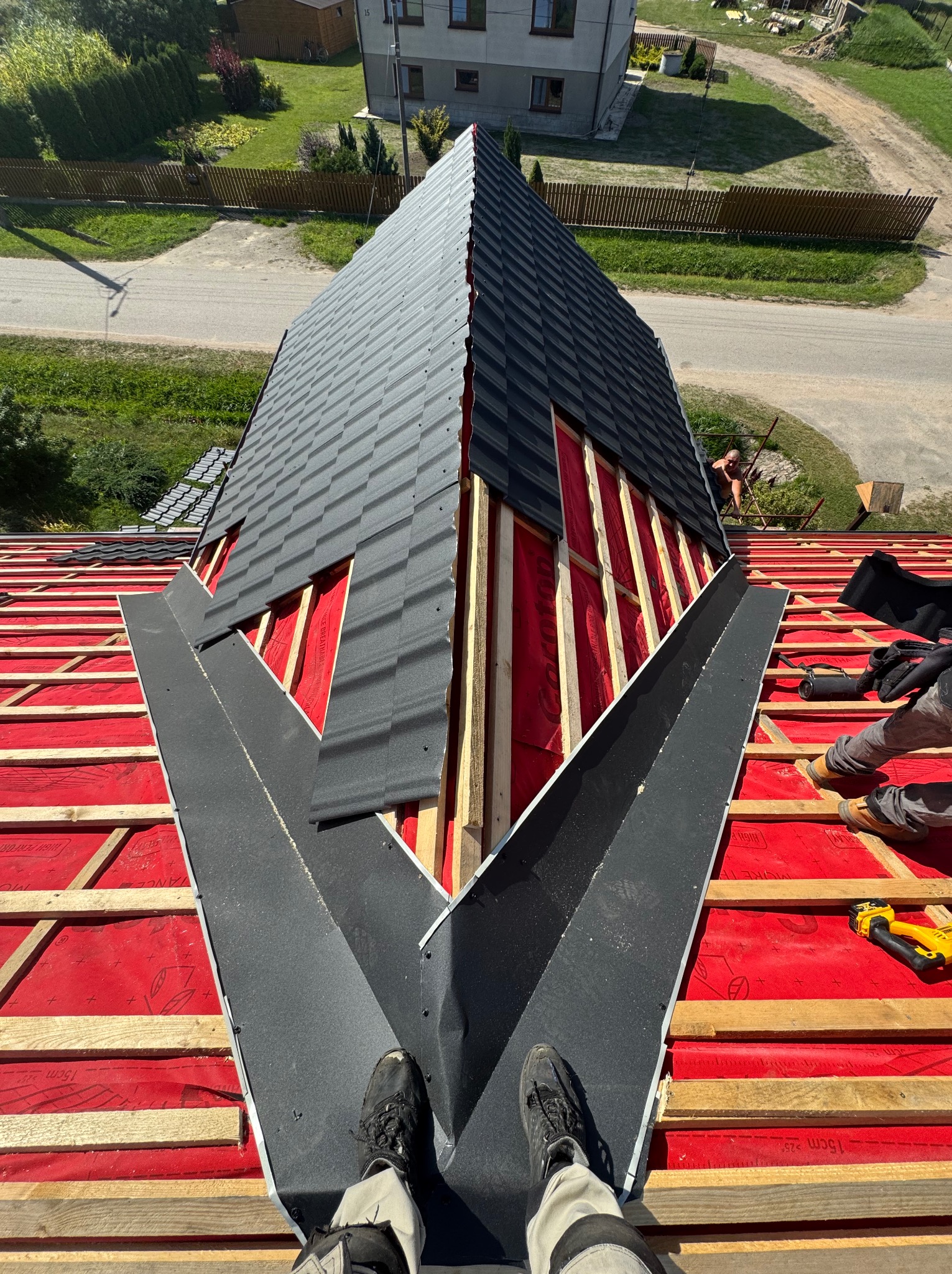 Montaż obróbek blacharskich na dachu z czerwoną membraną, widok z góry na fragment dachu z częściowo ułożonym pokryciem w kolorze grafitowym.