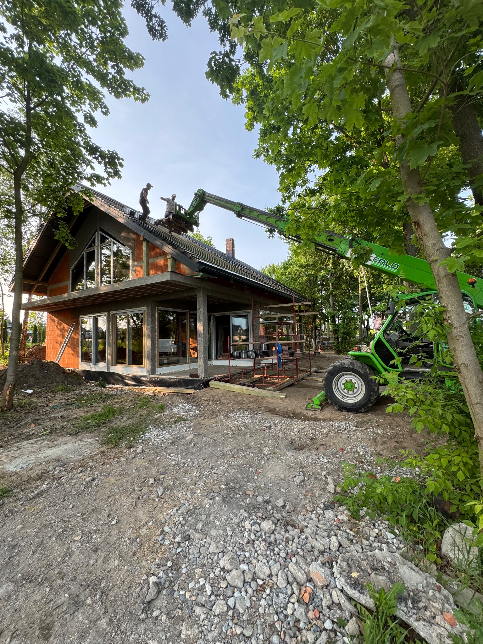 Montaż dachu na nowoczesnym domu jednorodzinnym z użyciem podnośnika teleskopowego. Praca na wysokości, widoczne elementy konstrukcyjne i ceglana elewacja.