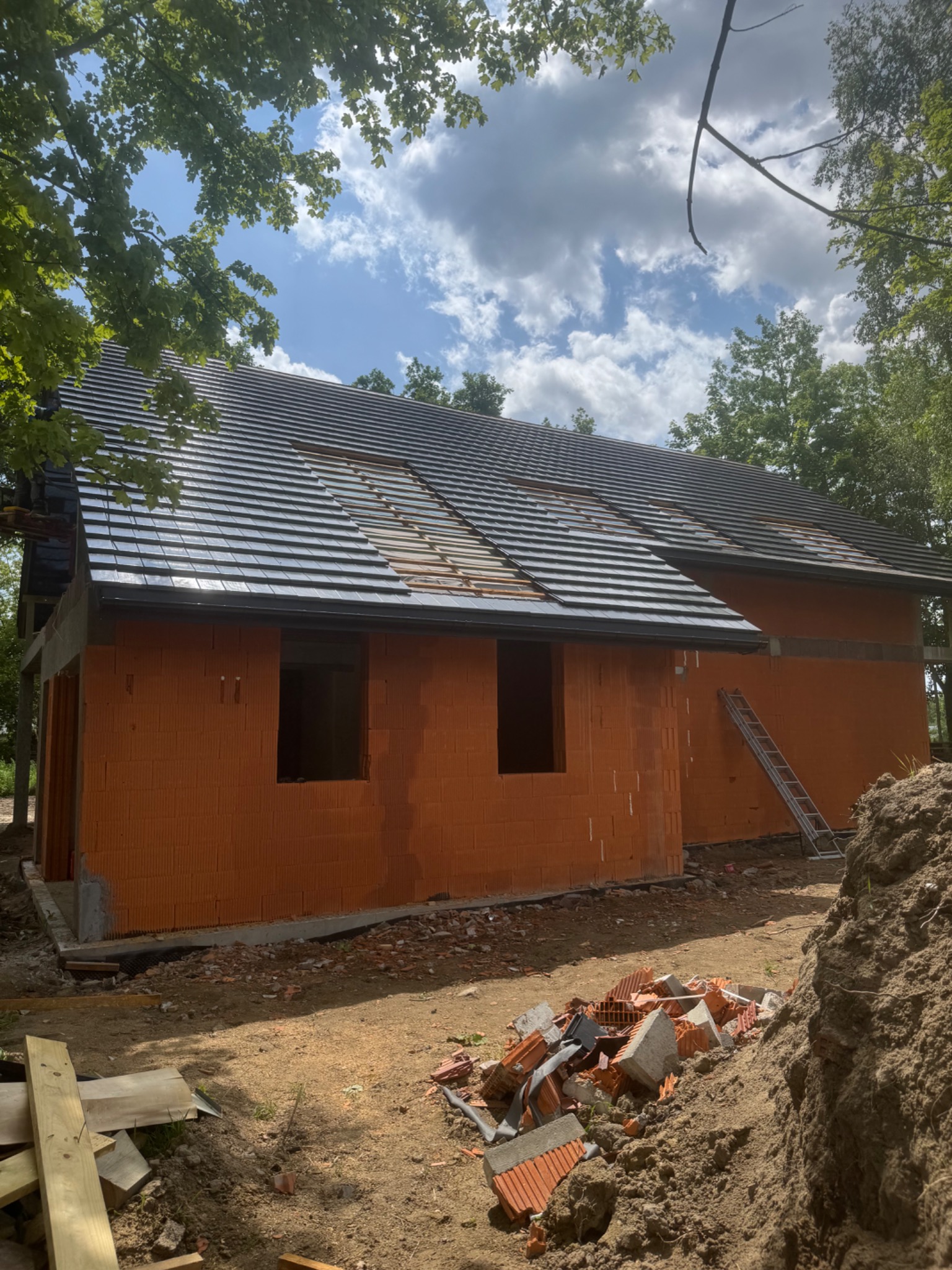 Budowa domu z częściowo pokrytym dachem dachówką ceramiczną. Widoczne ściany z pustaków ceramicznych i drabina oparta o ścianę. Prace w toku, teren budowy.