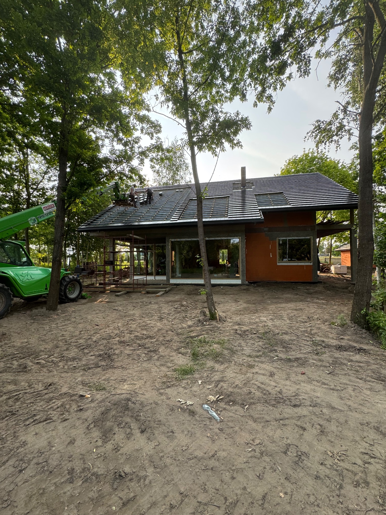 Montaż ciemnej dachówki na nowym domu z pomarańczową elewacją, widoczny z poziomu gruntu, z użyciem podnośnika teleskopowego na tle drzew.