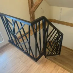 Czarna, geometryczna balustrada schodowa z metalu na drewnianych schodach, widok z góry, z elementami drewnianej konstrukcji stropu widocznymi w tle.