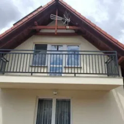 Czarna, metalowa balustrada balkonowa z pionowymi szczeblami na tle kremowej elewacji domu z brązowym dachem i anteną telewizyjną.